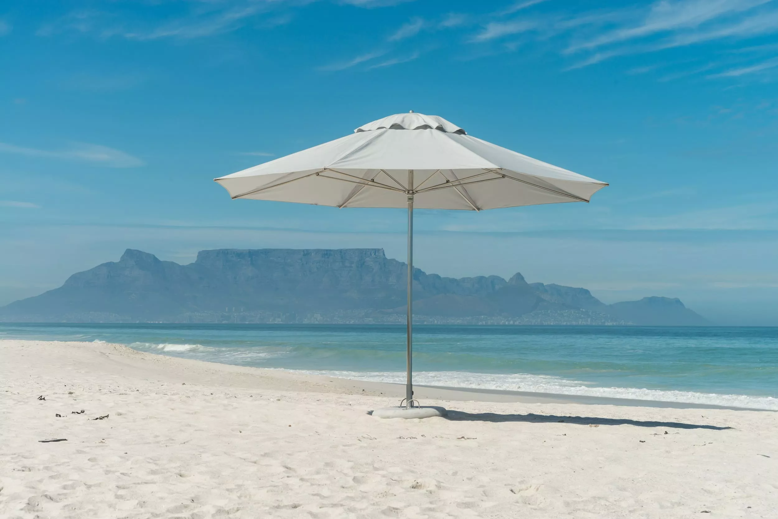 CapeUmbrellas ELITE Constantia Sonnenschirm in Weiß auf einem sandigen Strand mit Blick auf das Meer und einen Berg im Hintergrund.