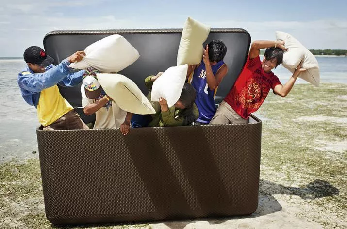 Fünf Personen machen eine Kissenschlacht in einer geöffneten Dedon La Malle Kissentruhe XL am Strand.