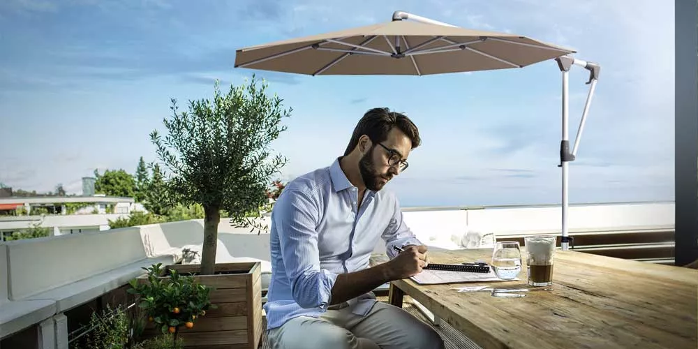 Ein Mann sitzt unter einem beigen Glatz Sunwing Casa Sonnenschirm auf einer Terrasse und arbeitet an einem Holztisch.
