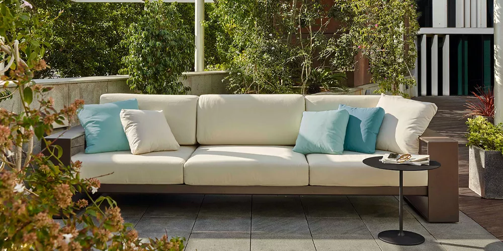 Andreu World Landscape Alu Sofa in Cremeweiß auf einer Terrasse mit Pflanzen im Hintergrund, begleitet von blauen Kissen und einem Beistelltisch.