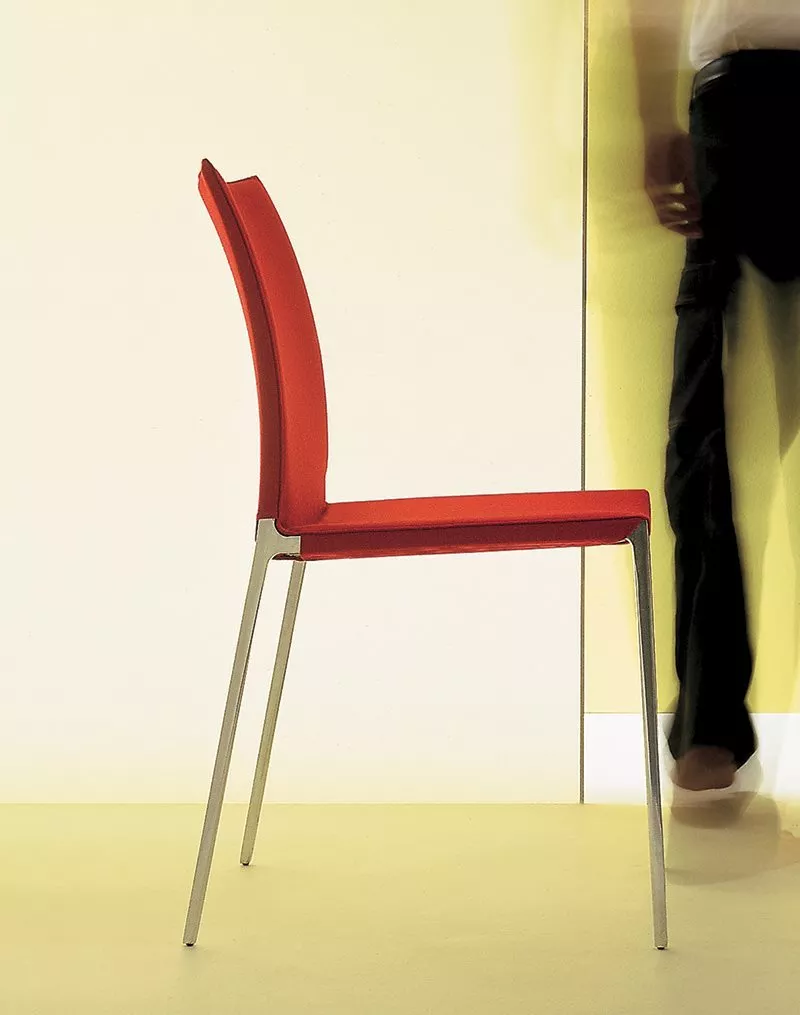 Ein roter Zanotta Lia Stuhl mit Metallbeinen steht in einem minimalistischen Raum während eine Person im Hintergrund vorbeigeht.