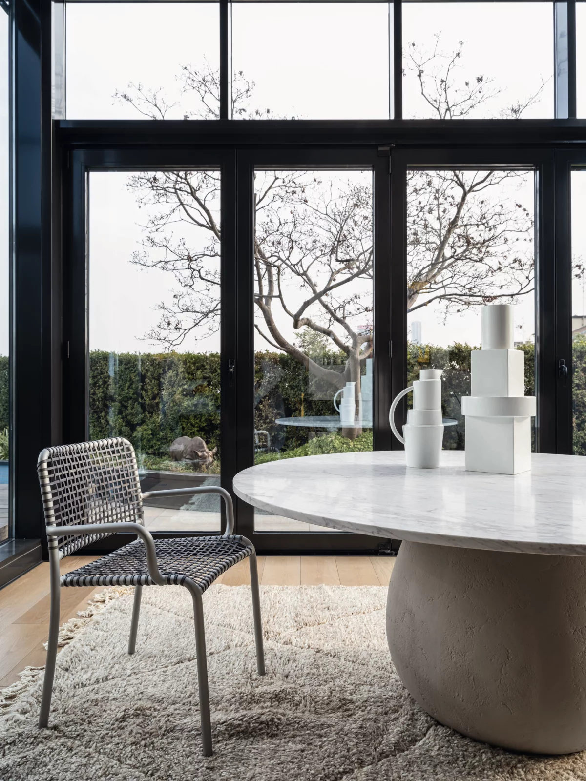 Ein Gervasoni Stuhl und ein runder Tisch mit Dekoration in einem modernen Raum mit großen Fenstern und Blick auf einen Garten