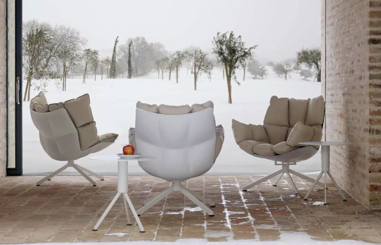Drei B&B Italia Husk Outdoor Loungesessel in verschiedenen Farben stehen auf einer Terrasse im Winter mit schneebedeckter Landschaft im Hintergrund.