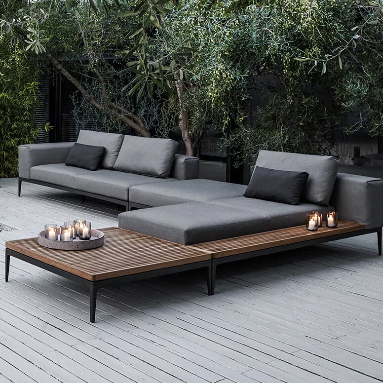 Lounge-Gartenmöbel Grid von Gloster in Grau auf einer Holzterrasse, umgeben von grünen Bäumen, mit Kerzen auf dem Tisch.