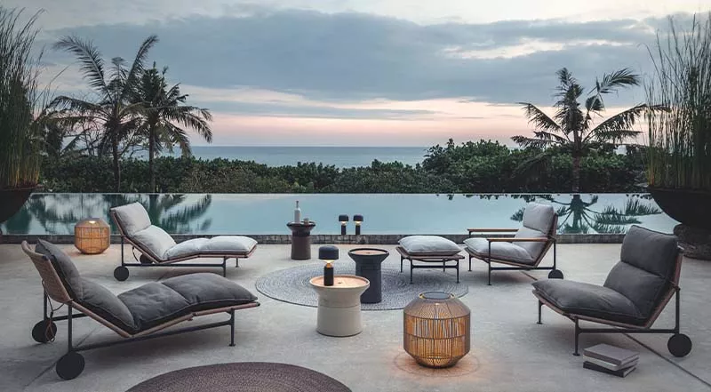 Gartenmöbel-Set Gloster Zenith in Grau auf einer Terrasse mit Meerblick bei Sonnenuntergang, bestehend aus Liegen und Sesseln.