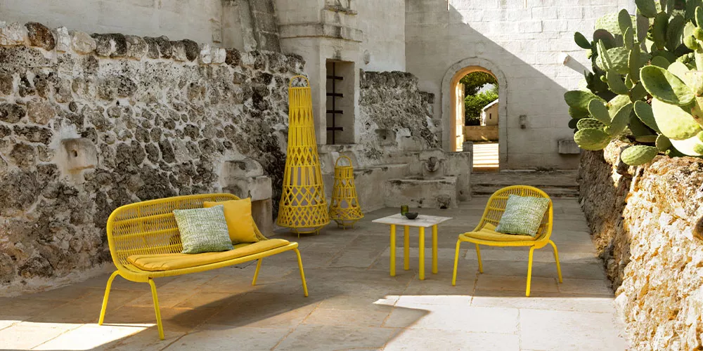 Gelbe Talenti Panama Kollektion Gartenmöbel auf Steinboden vor historischer Steinmauer mit Kakteen im Hintergrund.