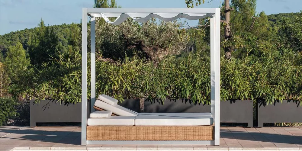 Point Sunset Outdoor Daybed in Weiß und Beige auf einer Terrasse mit grüner Pflanzenkulisse, ideal für entspannte Momente im Freien.