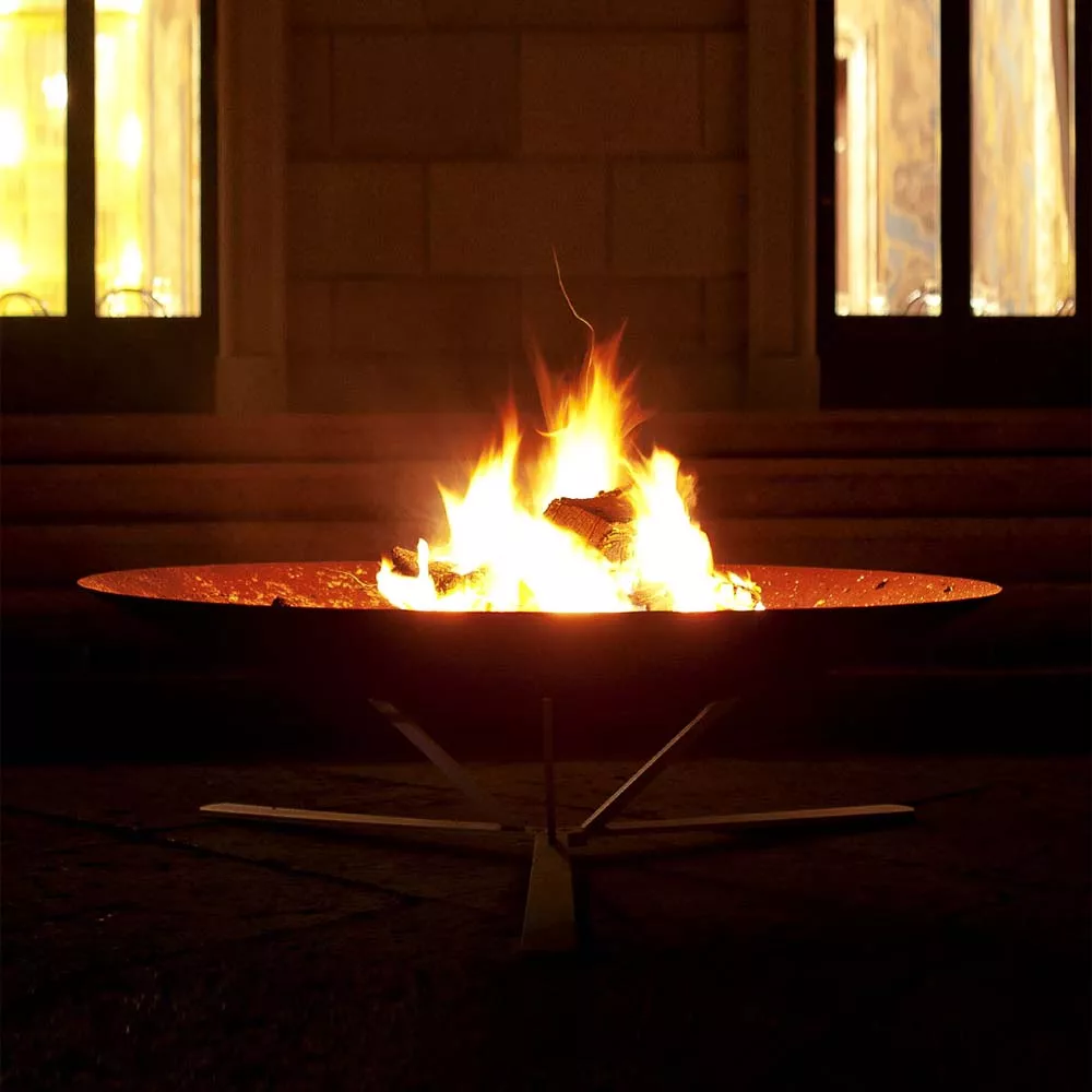 AK47 Discolo Feuerstelle aus oxidiertem Stahl 120 cm im Einsatz bei Nacht mit brennendem Feuer im Garten vor einem Gebäude