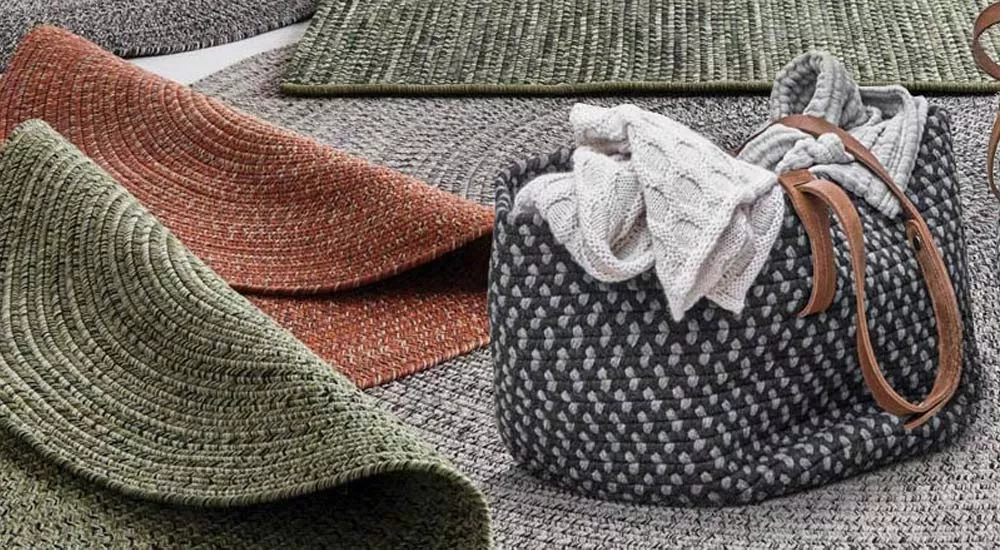 Gloster Teppiche in Grün, Rot und Grau, aus grobem Stoff, auf einem hellen Hintergrund, neben einem geflochtenen Korb mit Decken.