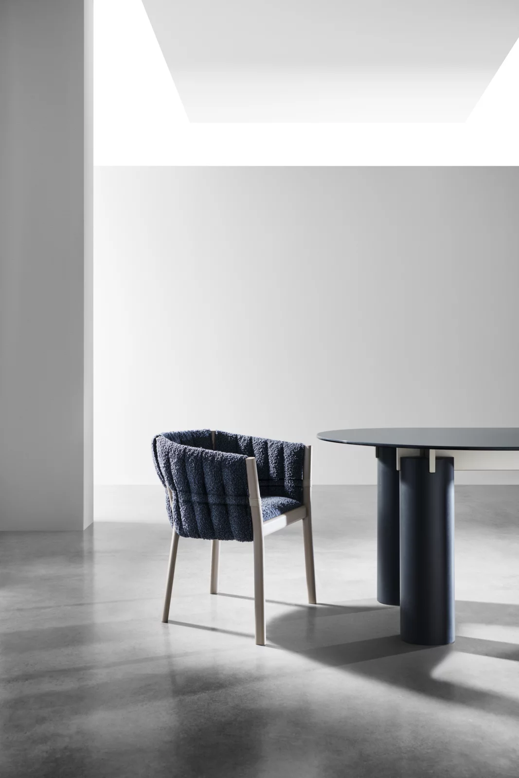 Gervasoni Inout 823 Sessel in dunkelblauem Stoff und hellem Holzrahmen neben einem modernen Tisch in einem minimalistischen Raum