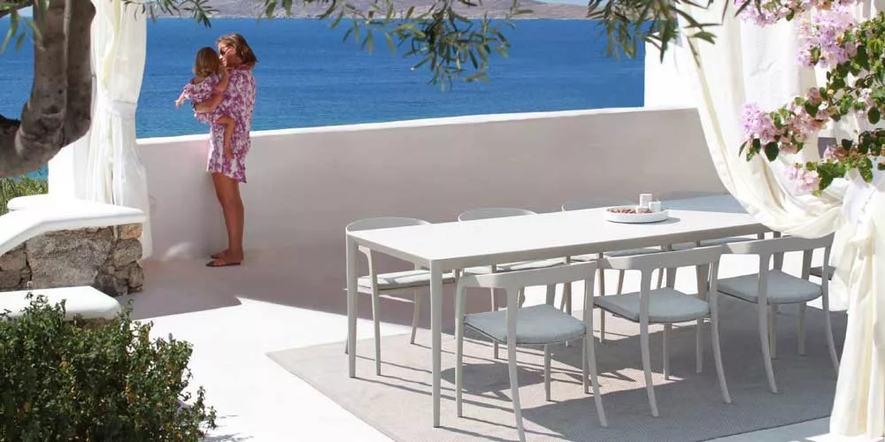 Royal Botania U-Nite Essgruppe in Weiß auf einer Terrasse mit Meerblick, umgeben von weißen Vorhängen und blühenden Pflanzen.