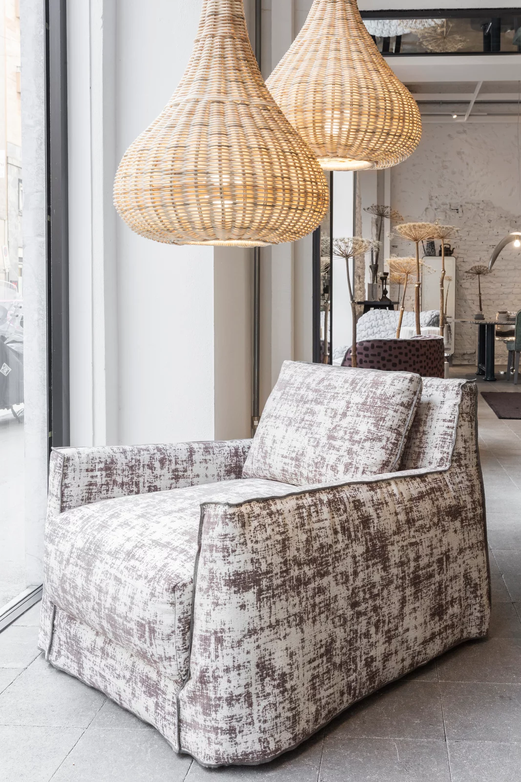 Ein gemütlicher Sessel von Gervasoni in einem modernen Showroom mit großen Fenstern und hängenden Rattanlampen im Hintergrund.