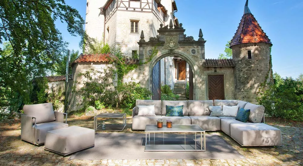 Fischer Möbel Suite Loungegruppe in Beige auf einer Terrasse vor einem historischen Gebäude, bestehend aus Sofa, Sessel und Tisch.