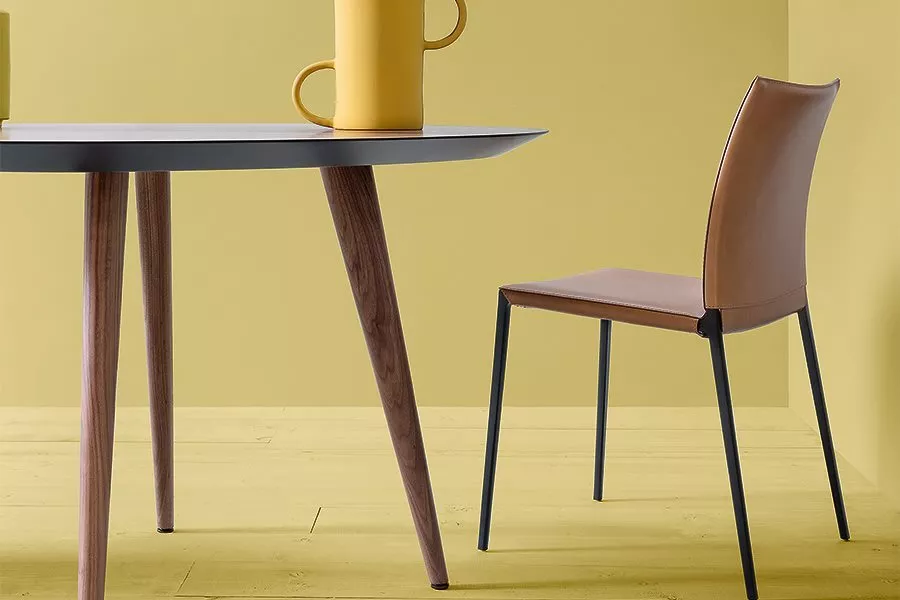 Braunlederstuhl vor gelbem Hintergrund neben einem Tisch mit Holzbeinen und gelber Kanne, moderne Inneneinrichtung, seitliche Ansicht.