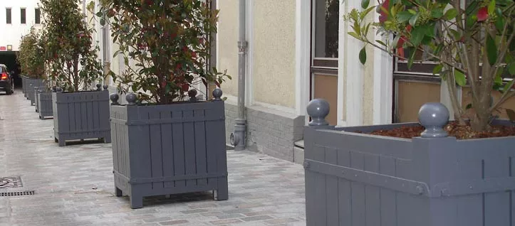 Mehrere Orangerie Pflanzkübel aus lackiertem Akazienholz stehen in einer Reihe auf einem gepflasterten Weg vor einem Gebäude.