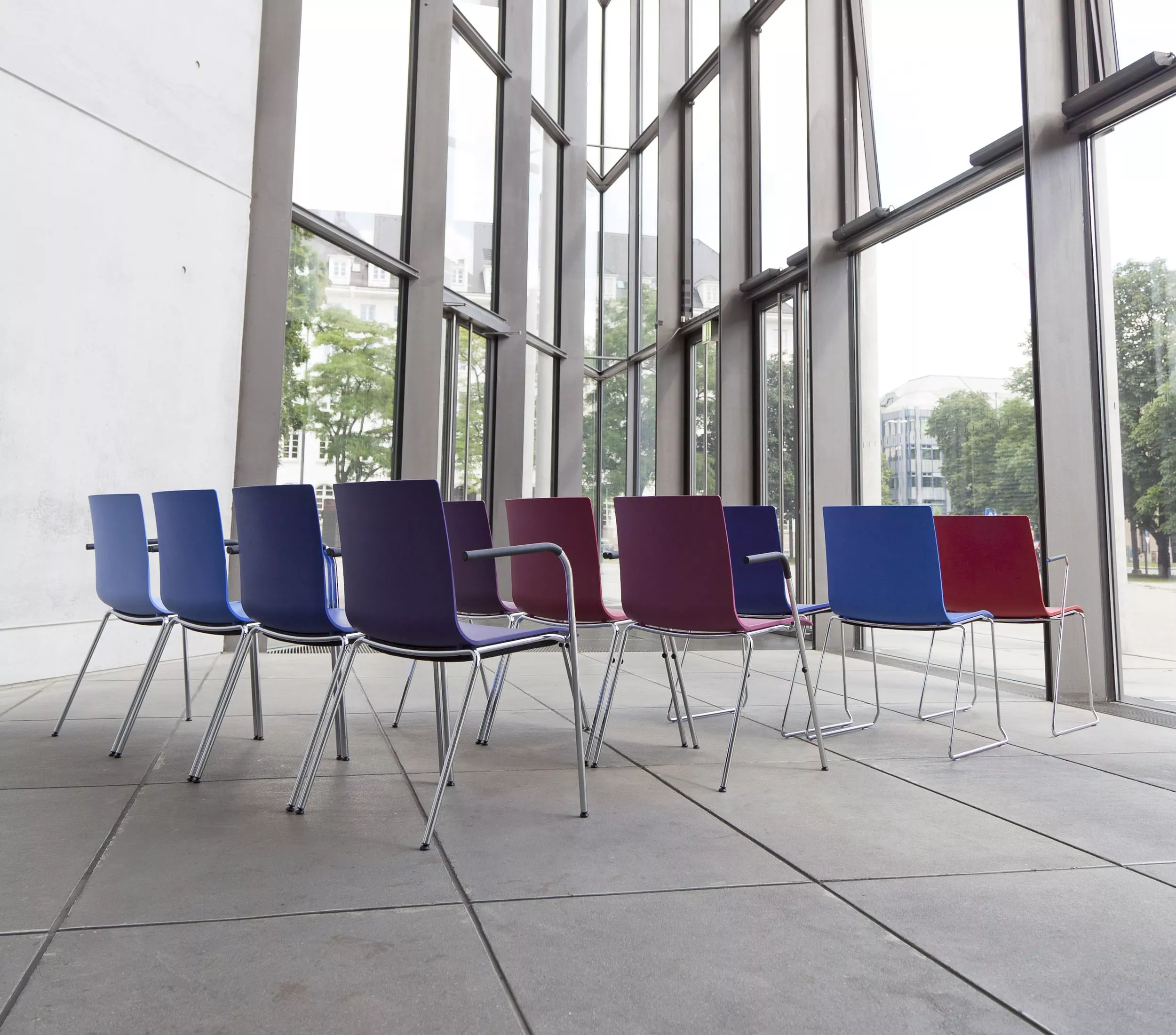 Mehrere Thonet S 160 F Stühle in verschiedenen Farben stehen in einem modernen Raum mit großen Fenstern und Blick auf eine städtische Umgebung