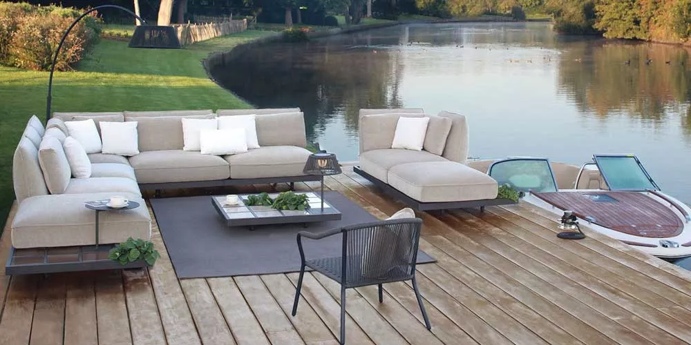 Royal Botania Mozaix Loungegruppe in Beige auf Holzterrasse am See, stimmungsvolles Outdoor-Setting mit Boot im Hintergrund.