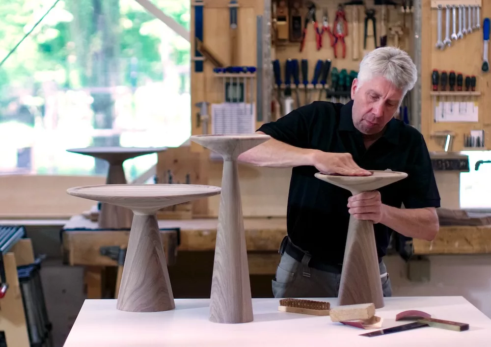 Ein Handwerker bearbeitet in einer Schreinerei zwei Figura Beistelltische von Draenert aus Holz im Hintergrund sind Werkzeuge und weitere Tische zu sehen