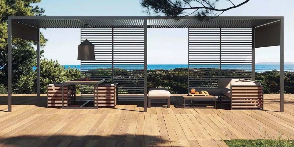 Gandia Blasco Pergolas in Anthrazit auf einer Holzterrasse mit Meerblick, moderne Outdoor-Möbel darunter, stimmungsvolle Landschaft.