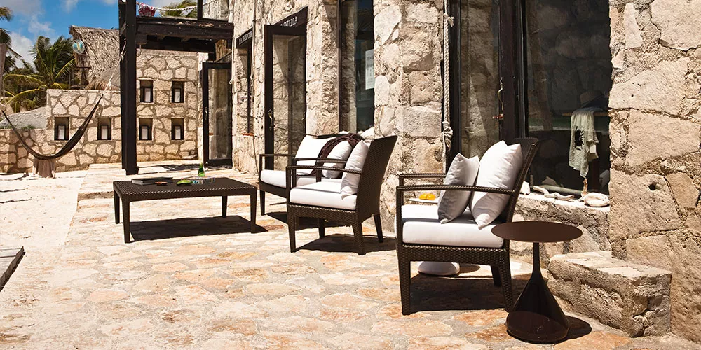 Dedon Kollektion Panama zeigt eine stilvolle Outdoor-Sitzgruppe mit zwei Sesseln einem Tisch und einem Beistelltisch vor einem Steinhaus in sonniger Umgebung