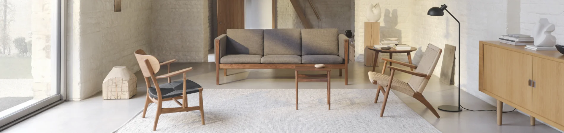 Carl Hansen Wohnzimmermöbel in einem modernen, hellen Raum mit Holzmöbeln und grauen Polstern auf einem Teppich vor einer Backsteinwand.