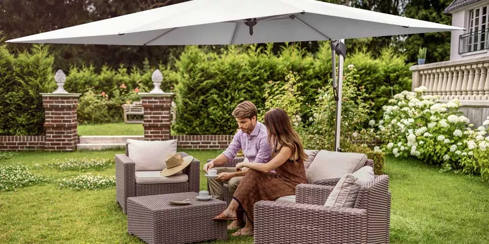 Ein Paar sitzt unter dem grauen Glatz Sombrano Sonnenschirm im Garten auf Rattanmöbeln, umgeben von grüner Hecke und Blumen.