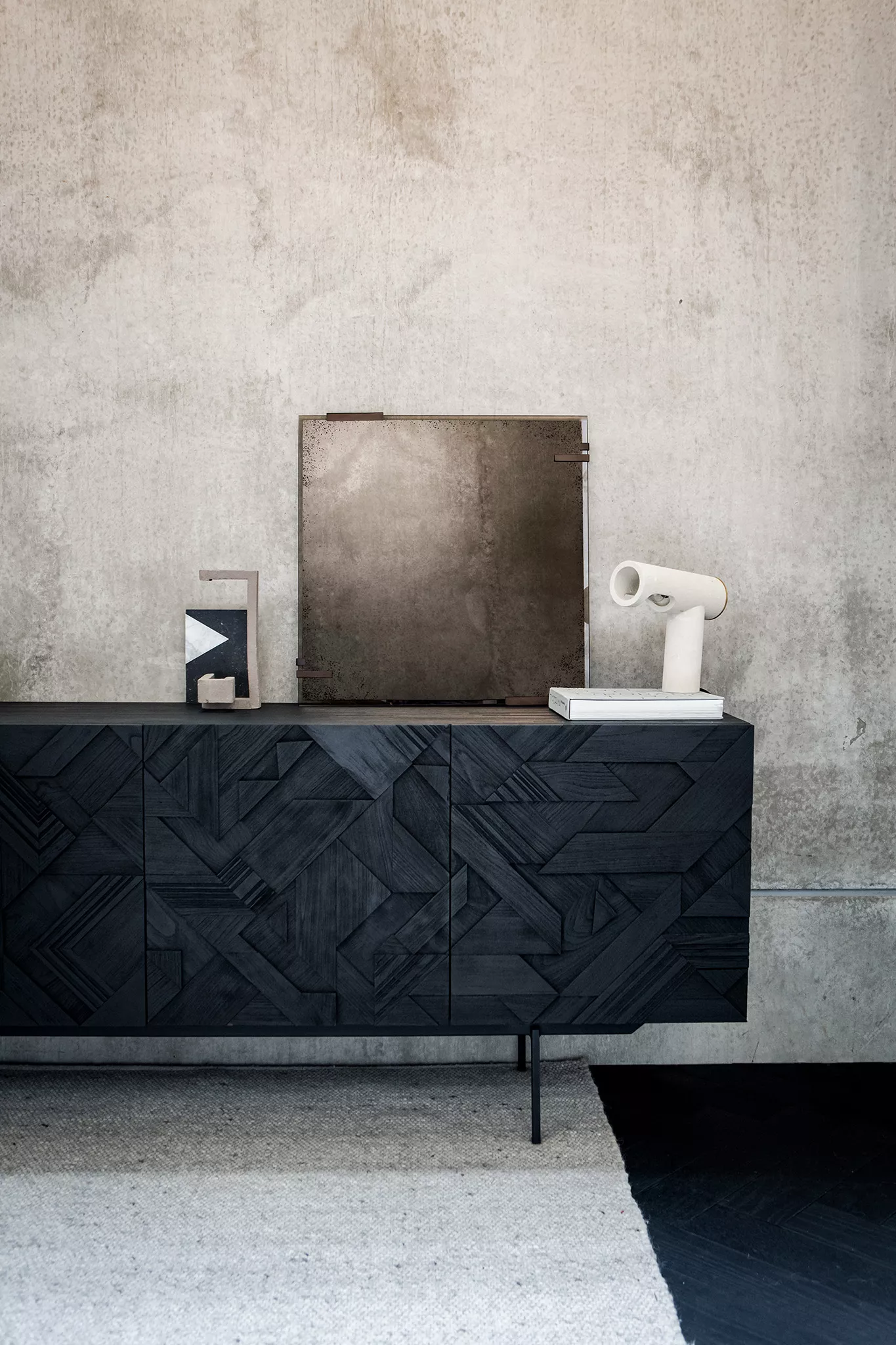 Bronzefarbener, rahmenloser Wandspiegel auf schwarzem, grafischem Sideboard mit vier Türen vor einer Betonwand.