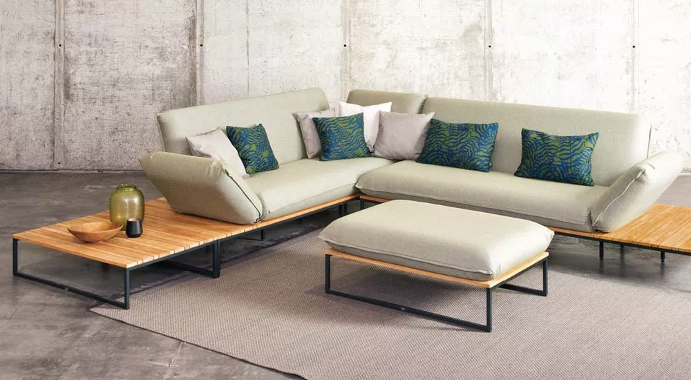 Fischer Möbel Flora Loungegruppe in Beige mit Holzelementen und grünen Kissen auf grauem Teppich vor Betonwand, Perspektive von vorne.