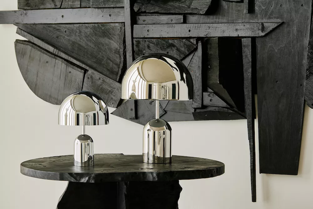 Tom Dixon Bell Tischleuchte in Chrom auf einem rustikalen Holztisch vor einer abstrakten schwarzen Wandkunst.