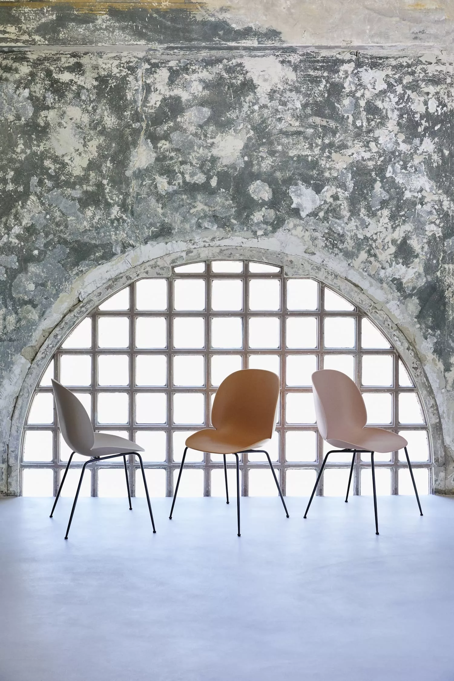 Drei Gubi Stühle in verschiedenen Farben stehen vor einer alten gemauerten Wand mit einem großen Fenster