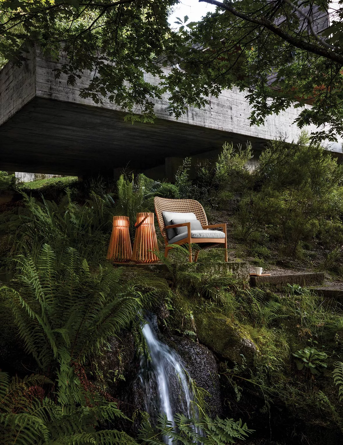 Gloster Kay Schaukelsessel in Naturfarbe auf moosigem Waldboden vor modernem Betongebäude, umgeben von Farnen und kleinem Wasserfall.