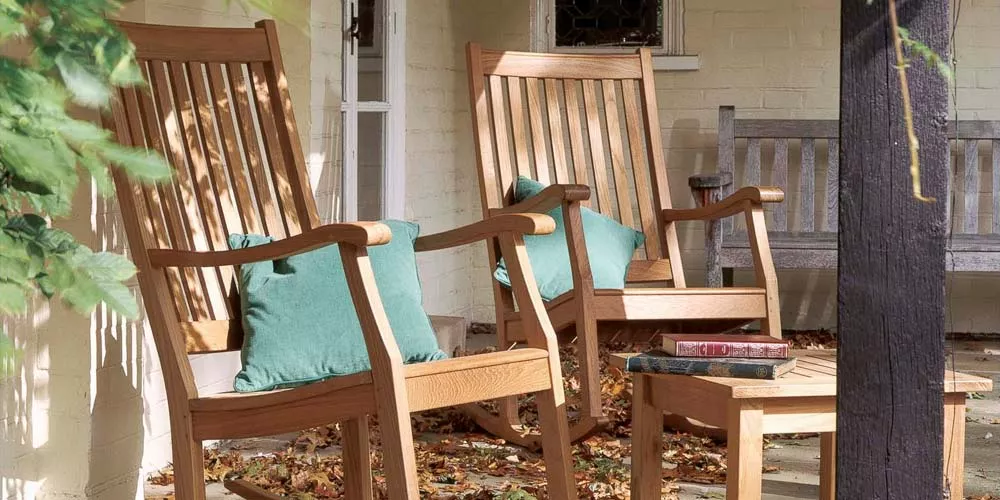 Zwei Barlow Tyrie Newport Holzstühle mit grünen Kissen auf einer Terrasse, umgeben von Herbstlaub, vor einer hellen Hauswand.
