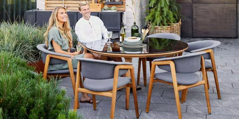 Cane-line Luna Essgruppe in Grau und Holzoptik auf einer Terrasse mit Pflanzen im Hintergrund, stimmungsvolle Outdoor-Szene.