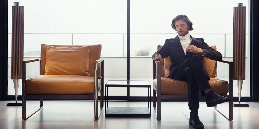 Ein Mann in einem eleganten Anzug sitzt auf einem Roshults Monaco Ledersessel in Cognac vor einer großen Fensterfront.