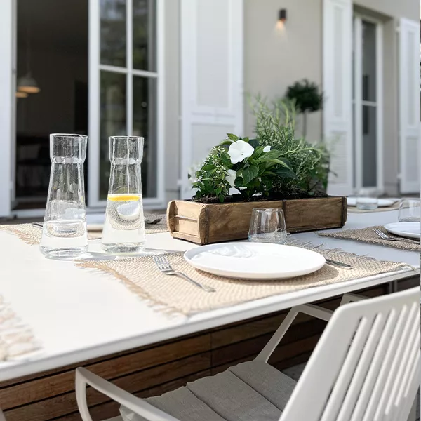Außenbereich einer Villa mit gedecktem Tisch, weißen Stühlen und Blumendekoration auf einer Terrasse bei Tageslicht.