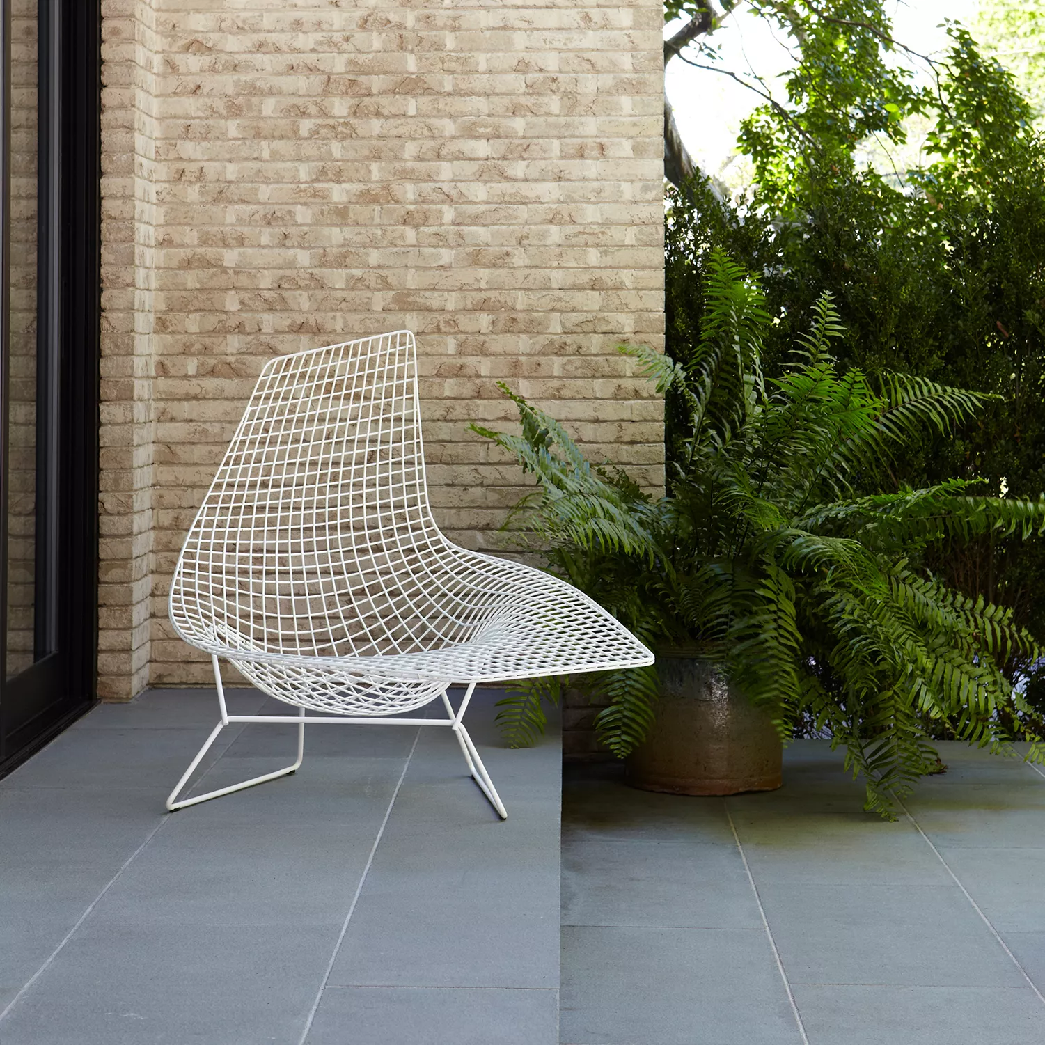 Knoll Harry Bertoia Outdoor Loungesessel in Weiß steht auf einer Terrasse neben einem großen Farn und einer Backsteinmauer im Hintergrund