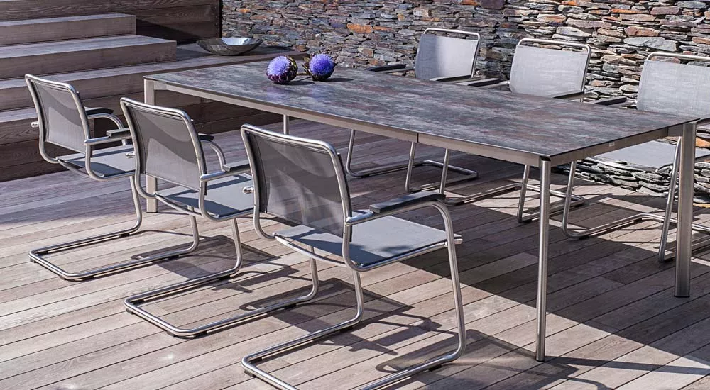 Fischer Möbel Swing Essgruppe in Grau auf Holzterrasse vor Steinmauer, bestehend aus großem Tisch und sechs Stühlen, in moderner Umgebung.