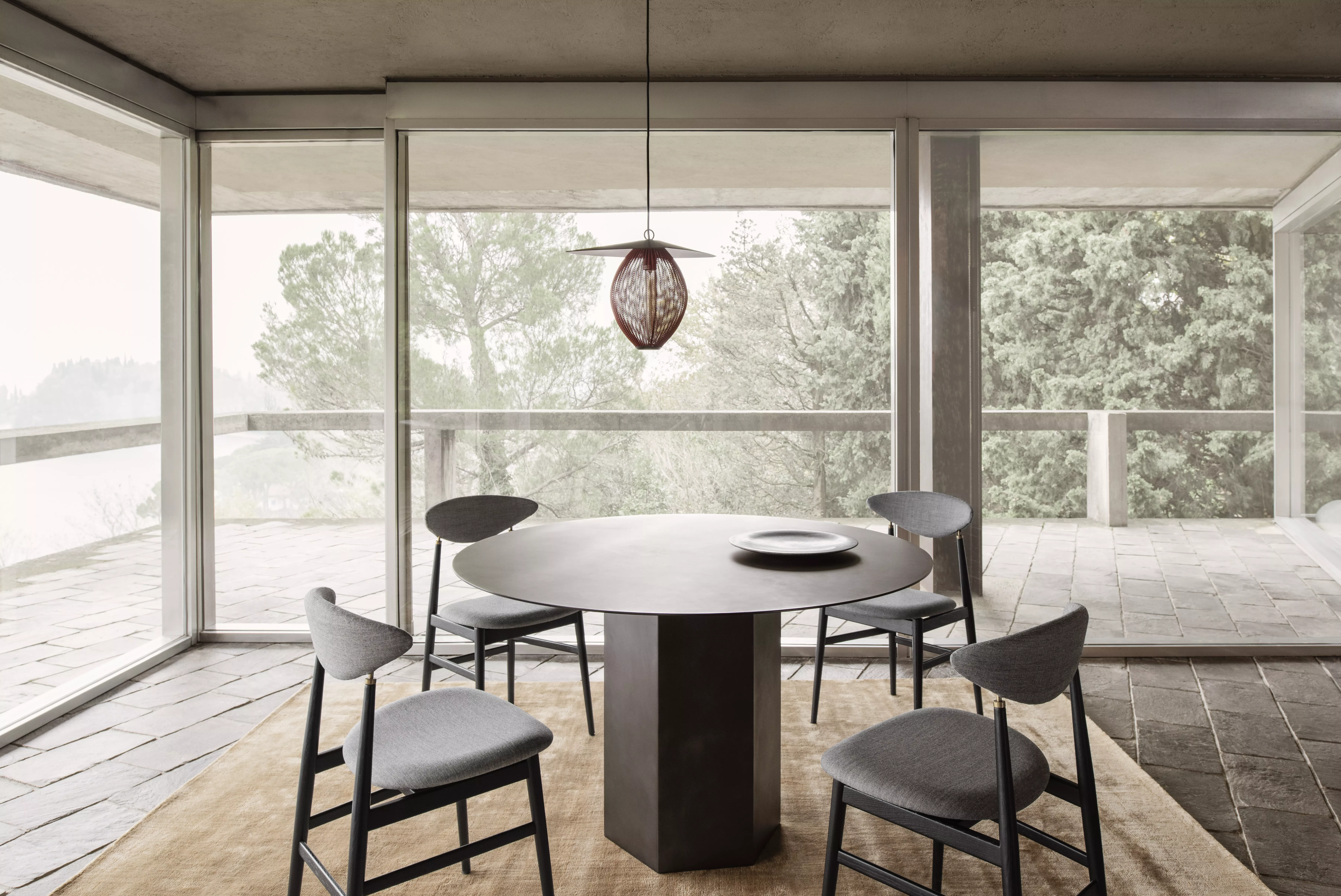 Ein eleganter Essbereich mit einem runden Gubi-Tisch und vier Stühlen in einem modernen Raum mit großen Fenstern und Blick auf die Natur.