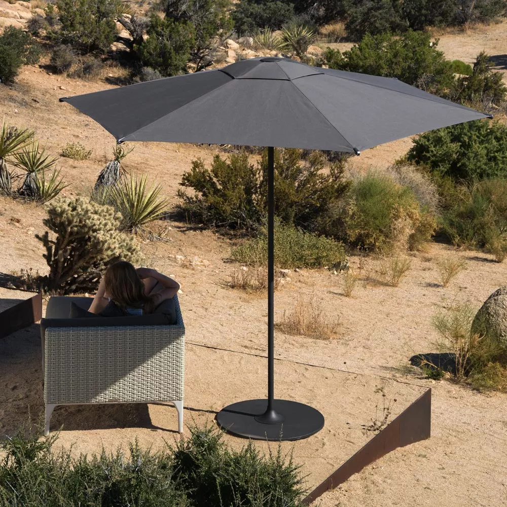 Dedon Reav Hexagon S Sonnenschirm in einer Wüstenlandschaft mit einer Frau die auf einem Sofa im Schatten des Schirms entspannt