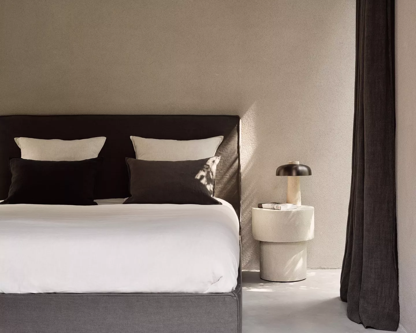 Gemütliches Schlafzimmer mit grauem Bett, weißen und schwarzen Kissen, beiger Wand und modernem Nachttisch mit Lampe, Tageslicht.