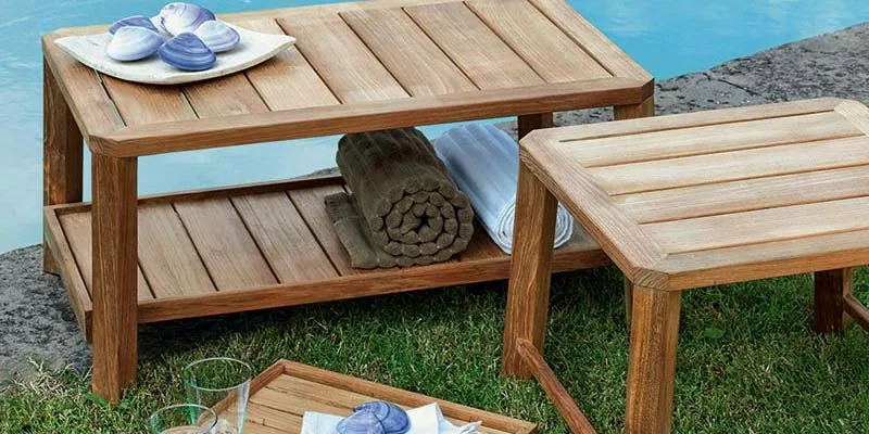 Ethimo Petit Club Outdoor Tisch und Hocker aus Teakholz, aufgestellt auf einer Wiese neben einem Pool, mit Handtüchern und Gläsern dekoriert.