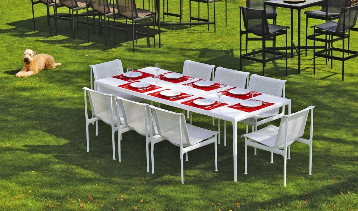 Knoll Studio 1966 Collection weißer Esstisch mit acht Stühlen auf grünem Rasen gedeckt mit roten Platzsets und Geschirr im Hintergrund ein Hund