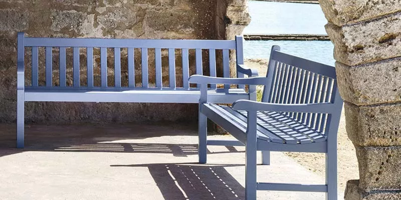 Ethimo Nothing Hill Bank in Blau, aus Holz, auf einer sonnigen Terrasse mit Steinmauer im Hintergrund.