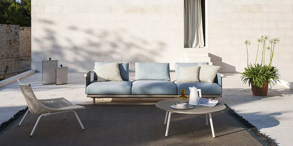 Roda Kollektion Eden, hellblaues Sofa auf einer Terrasse mit hellem Steinboden, begleitet von einem runden Tisch und einem Stuhl.