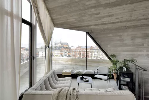 Gemütliches Wohnzimmer mit grauem Sofa, zwei Sesseln und Pflanzen, mit Blick auf die Stadt durch große Fenster unter einer schrägen Decke.