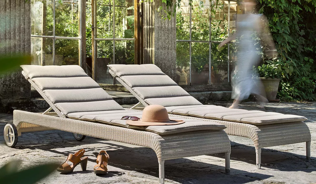 Zwei beige Vincent Sheppard Liegen auf einer Steinterrasse vor einem verglasten Gebäude, mit Strohhut und Sandalen im Vordergrund.
