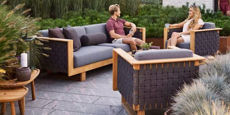 Zwei Personen entspannen auf der grauen Cane-line Angle Loungegruppe aus Holz in einem Garten mit grüner Pflanzenkulisse.