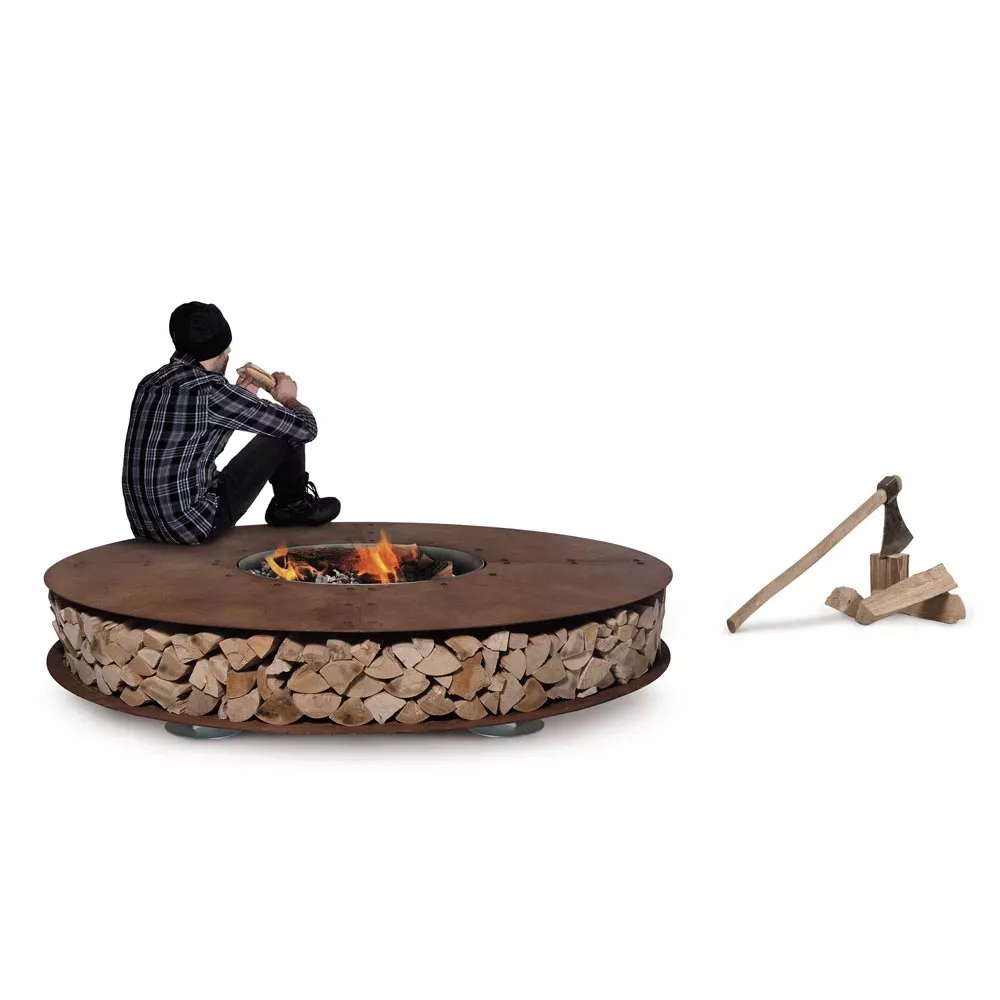 Ein Mann sitzt auf der AK47 Zero Feuerstelle 250 cm Gartenkamin und liest ein Buch während ein Beil und Holz daneben liegen.