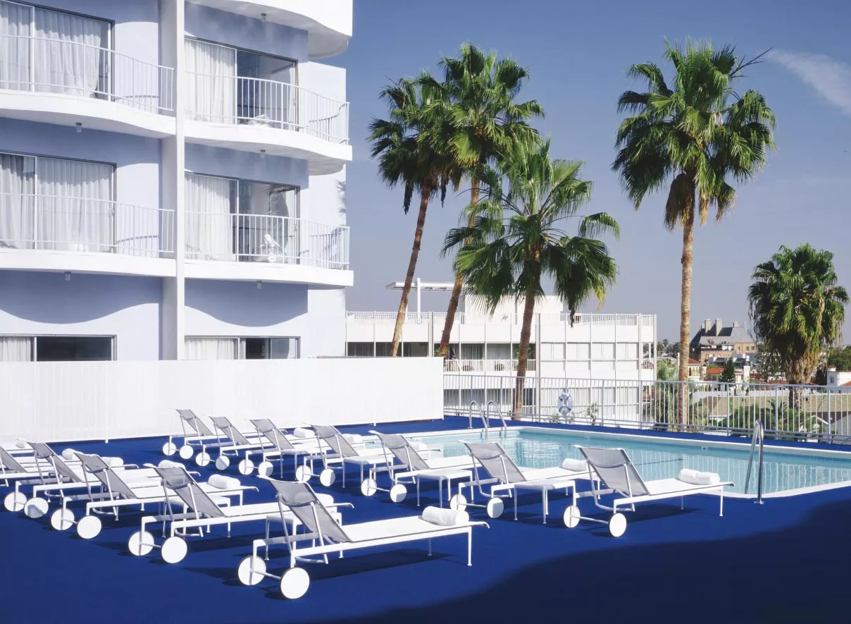 Knoll Studio 1966 Collection Sonnenliege verstellbar und rollbar auf einer blauen Terrasse neben einem Pool mit Palmen und einem weißen Gebäude im Hintergrund