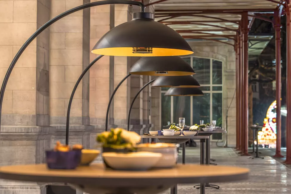 Heatsail Dome Arc Stehlampe in Schwarz, stimmungsvolle Beleuchtung auf einer überdachten Terrasse mit gedeckten Tischen.